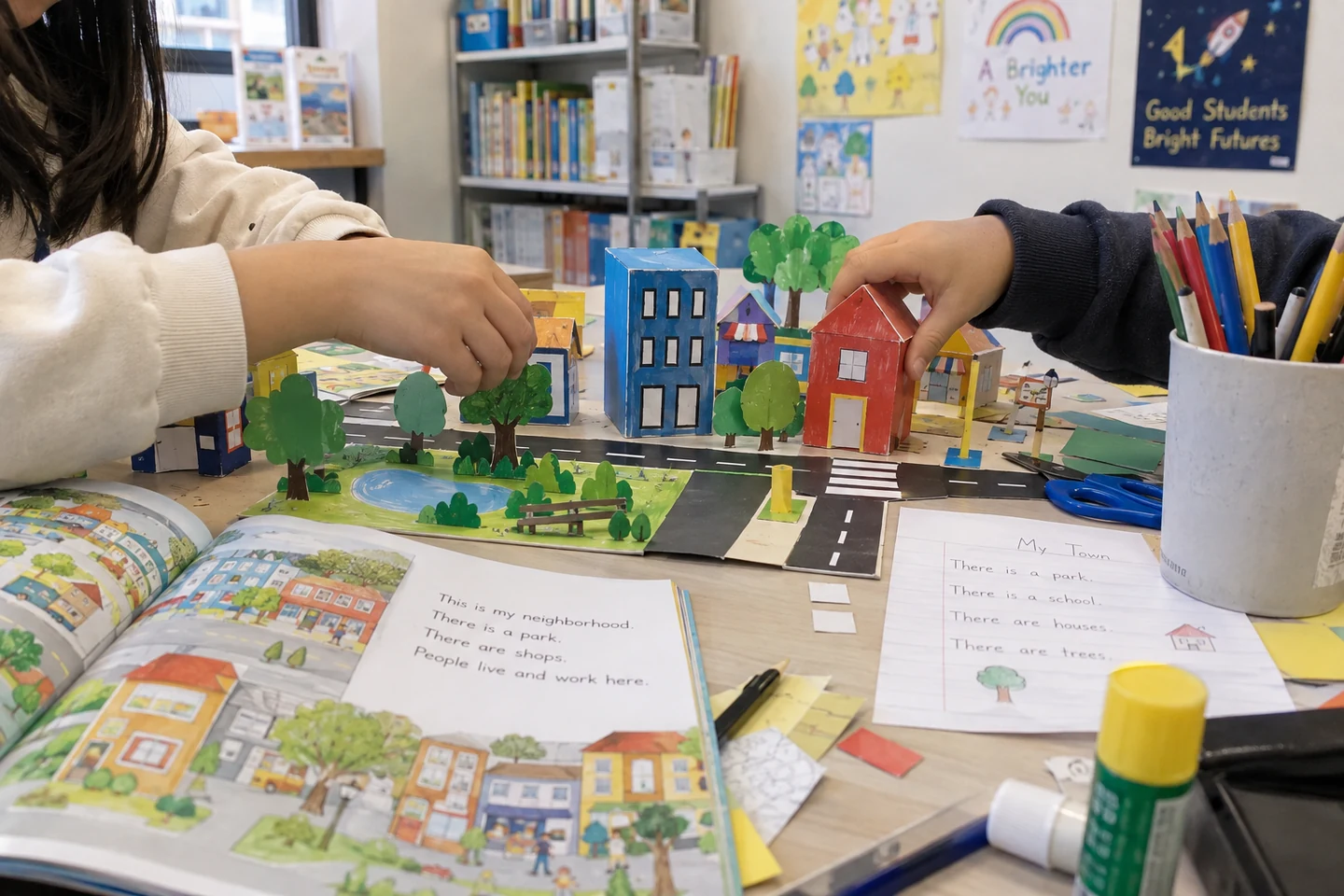 Students arrange handmade cardboard buildings beside an English reader and written project notes.